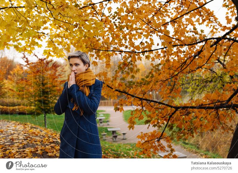 Kurzhaarige Frau wärmt im Herbst im Park ihre Hände mit ihrem Atem fallen Blätter kühl Ahorn umgefallene Blätter Herbstlaub Ahornblatt Laubwerk Herbstblattfarbe