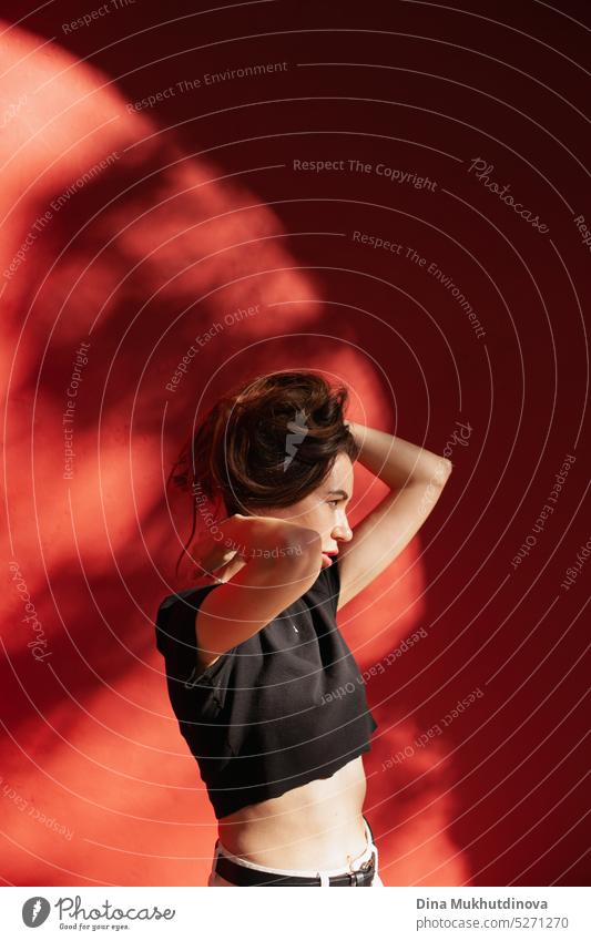 Junge Frau steht in der Nähe einer roten Wand und frisiert sich die Haare. Künstlerisches Foto mit weiblichem Modell und Schatten an der Wand. tausendjährig