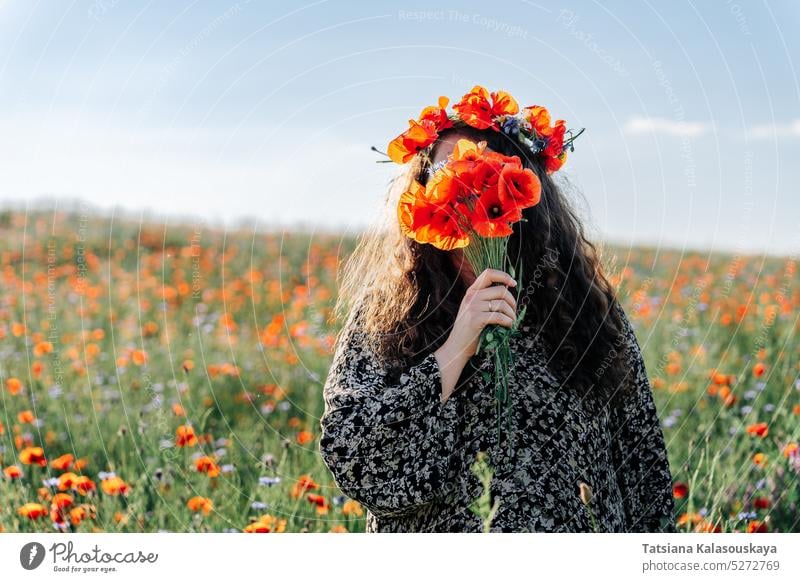 Langhaarige lockige Frau in Übergröße mit einem Kranz aus rotem Mohn steht inmitten eines Wiesenblumenfeldes und bedeckt ihr Gesicht mit einem Mohnblumenstrauß