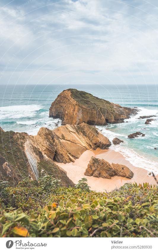 Steile Klippen, die in den gelb-orangen Sand eintauchen, der von den Wellen des Atlantiks in der Region Odemira, Portugal, bewässert wird. Auf den Spuren des Fischerpfads, Rota Vicentina