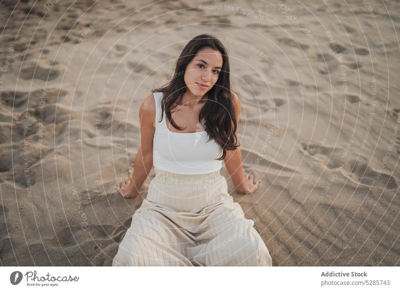 Unbeschwerte junge ethnische Frau am Strand sitzend bei Sonnenuntergang Lächeln Sand Feiertag Beine gekreuzt Kälte Tourist Glück positiv Sommer Urlaub