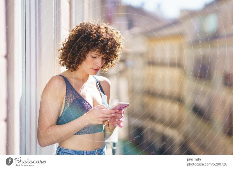 Junge Frau in BH und Jeans mit lockigem Haar beim Surfen auf dem Smartphone krause Haare Unterwäsche Frisur positiv Dessous Balkon Sommer ruhen Sonne heimwärts