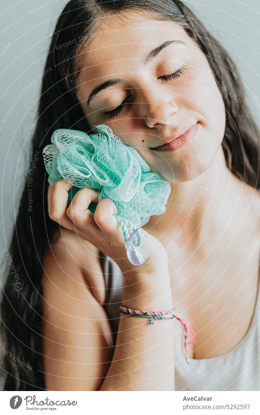 Fröhliche Frau, die einen Peeling-Schwamm hält und benutzt, während sie ihr Gesicht ausruht. Junge Erwachsene waschen ihren Körper mit Bad Luffa. Waschlappen Körper schrubben, closeup Hände reiben Haut im Bad. Persönliche Hygiene Konzept.