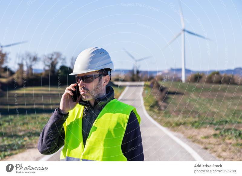 Ingenieur mit Schutzhelm spricht per Smartphone gegen Windkraftanlagen Mann Landschaft Fahrbahn Arbeiter benutzend Telefonanruf Turbine Öko alternativ Energie