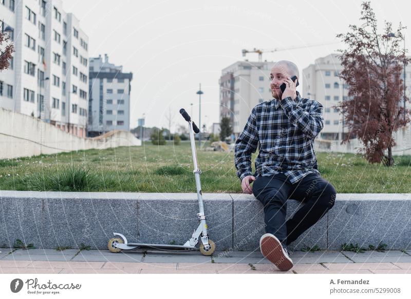 Junger Mann sitzt neben einem Motorroller und telefoniert in einem Park Tretroller Kickroller urban elektrisch Mitfahrgelegenheit Lifestyle modern Verkehr