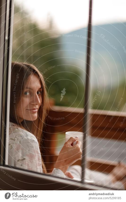 Junge Frau stehend nach dem Duschen am Morgen auf dem Balkon des Hotels. hält eine Tasse Kaffee oder Tee in ihren Händen. Blick nach draußen Natur Wald und Berg