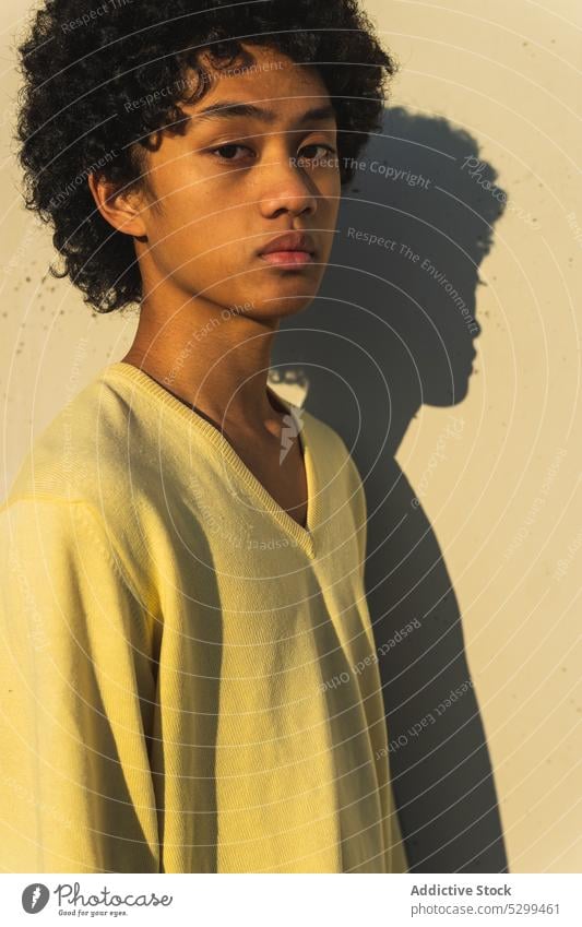 Ernster junger asiatischer Mann, der im Sonnenlicht in die Kamera schaut selbstbewusst cool Schatten Porträt gelb Vorschein Afro-Look Wand Stil ethnisch Frisur