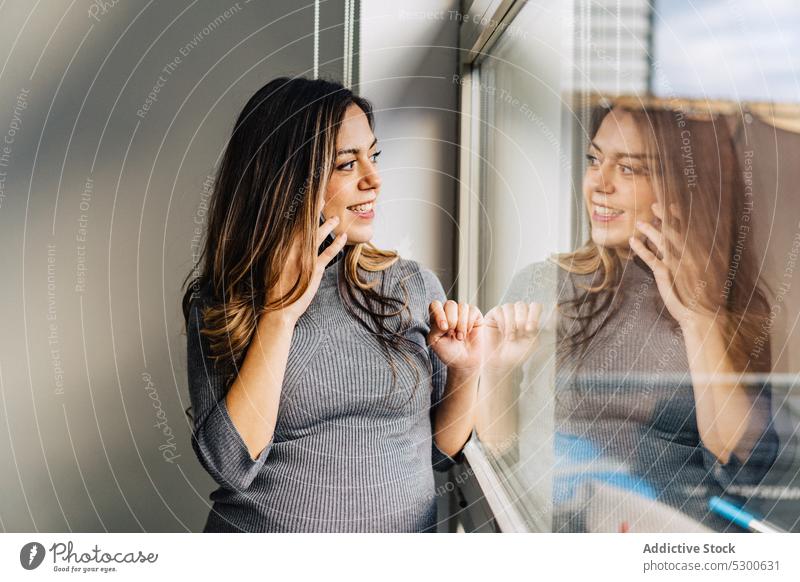 Lächelnde schwangere Frau, die mit einem Handy telefoniert benutzend Smartphone sprechen Fenster Telefonanruf Appartement Gespräch Kommunizieren positiv jung