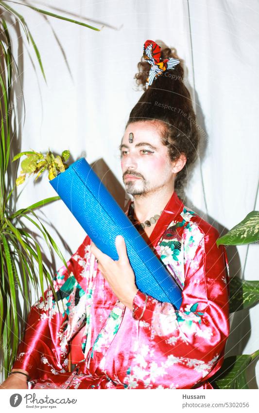 ist ein Fotokonzept über einen Mann, der eine Yogamatte hält, ein geblümtes Kleid trägt und seine Haare auf eine sehr lustige Weise gestylt hat. Sein Make-up sieht aus wie eine Geisha mit einem ironischen Blick.