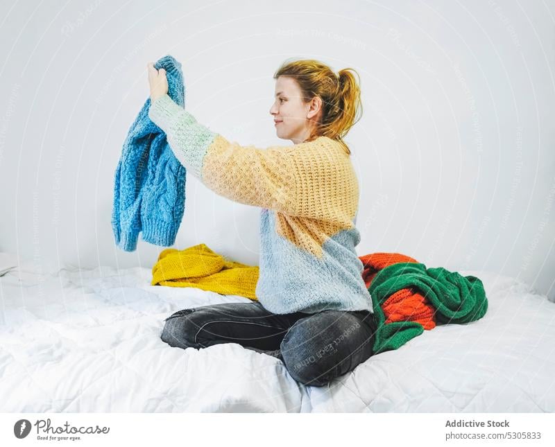 Glückliche Frau wählt Pullover auf dem Bett aus wählen Lächeln Komfort positiv heimwärts gemütlich jung Schlafzimmer weich Appartement Freude sitzen Inhalt