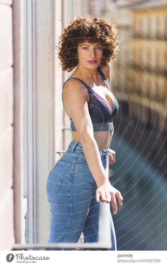 Junge Frau in BH und Jeans mit lockigem Haar auf sonnigem Balkon in die Kamera blickend heiter Sommer Glück krause Haare tagsüber Terrasse positiv Jeanshose