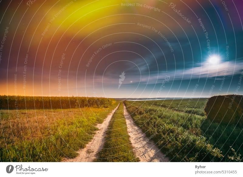 Night Starry Sky Above Country Straße durch Feld Wiese mit Heuballen nach der Ernte. Glühende Sterne und Vollmond über ländliche Landschaft. Bunte Fackel am Himmel. Landwirtschaftliche Landschaft unter Sternenhimmel. Bunte helle Flecken von Licht glühen in den Himmel