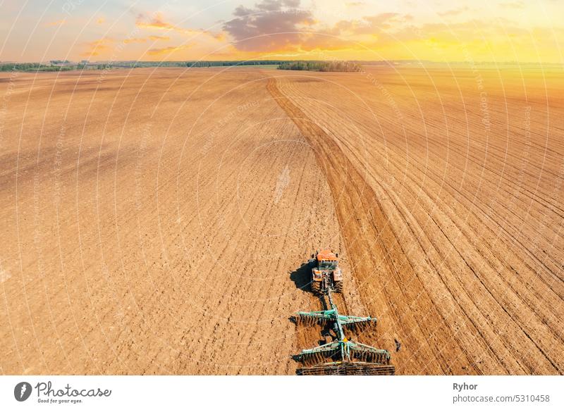 Erhöhte Ansicht Traktor Pflügen Feld im Frühjahr Saison. Beginn der landwirtschaftlichen Frühjahrssaison. Cultivator Pulled By A Tractor In Countryside Rural Field Landscape. Geänderte Sonnenuntergang Himmel.