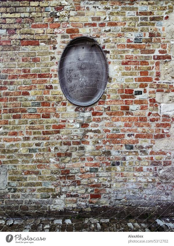 Sehr alte, verwitterte Backsteinwand mit einem verblichenem, ovalen, grauen Grabstein in der Mitte sehr alt Backsteine Mauer Wand Fassade Backsteinfassade