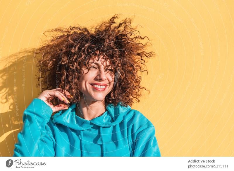 Charmante lockige Frau berührt Haar gegen gelbe Wand Porträt krause Haare Stil Individualität Farbe heiter Haare berühren charmant Outfit froh Lächeln Vorschein