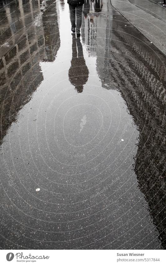 Eine Person (halb) mit Spiegelung (ganz) läuft auf einer nassen Strasse durch die Häuserschluchten einer Grossstadt. nach dem regen Spiegelungen im Wasser