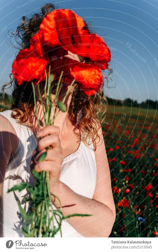 Selfie einer lockigen rothaarigen Frau, die ihr Gesicht mit einem Strauß roter Klatschmohnblüten inmitten eines Mohnfeldes bedeckt. Mohnblumen Wildblumen