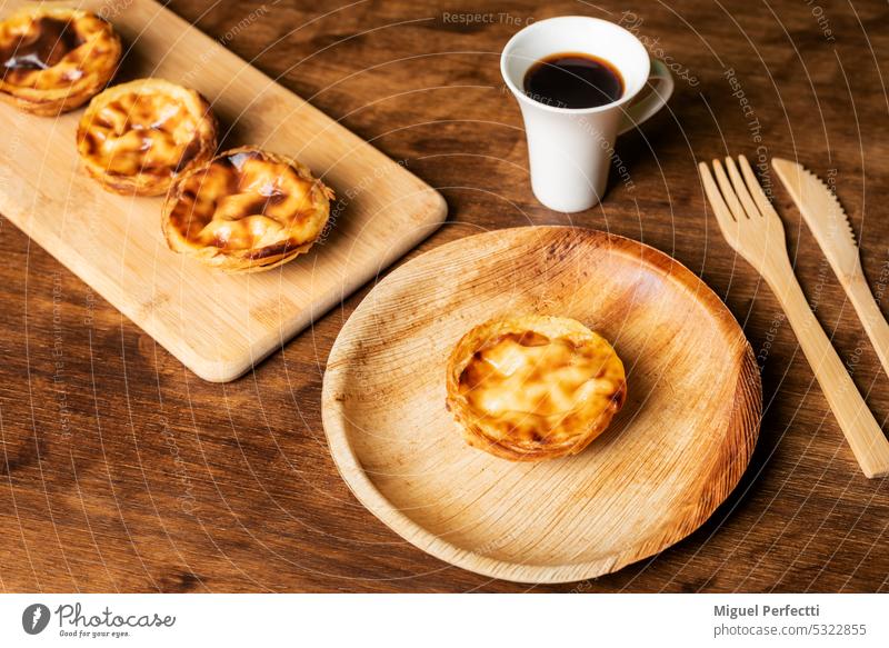 Pasteis de nata, eine typische Süßspeise aus Lissabon, Portugal, ein Kuchen auf einem Teller neben einer Tasse Kaffee, im Hintergrund mehrere weitere Kuchen.