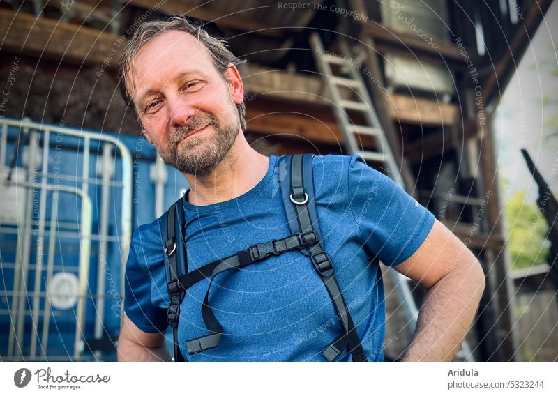 Mann im blauen T-Shirt und mit Rucksack schaut freundlich in die Kamera, Lagerschuppen/Werkstatt im Hintergrund Porträt Gesicht Lächeln Mensch Blick Bart