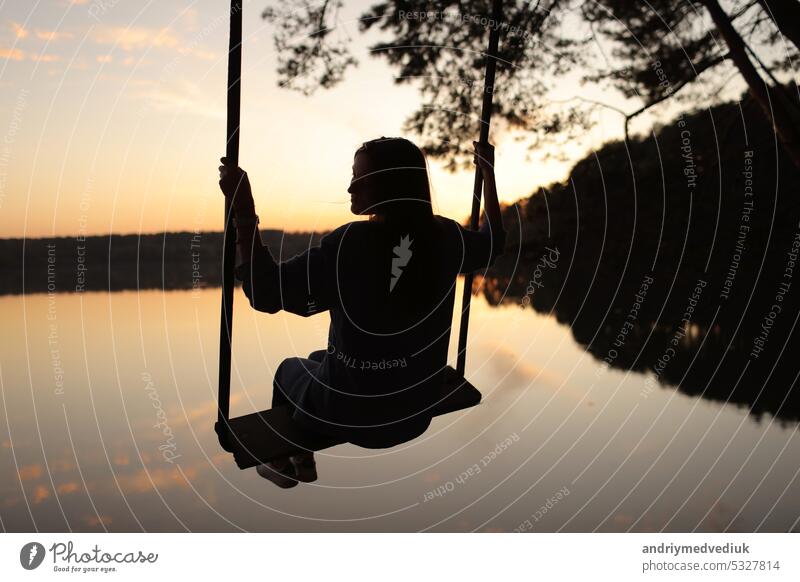 Silhouette einer romantischen jungen Frau auf einer Schaukel über einem See bei Sonnenuntergang. Junges Mädchen Reisende sitzen auf der Schaukel in der schönen Natur, Blick auf den See