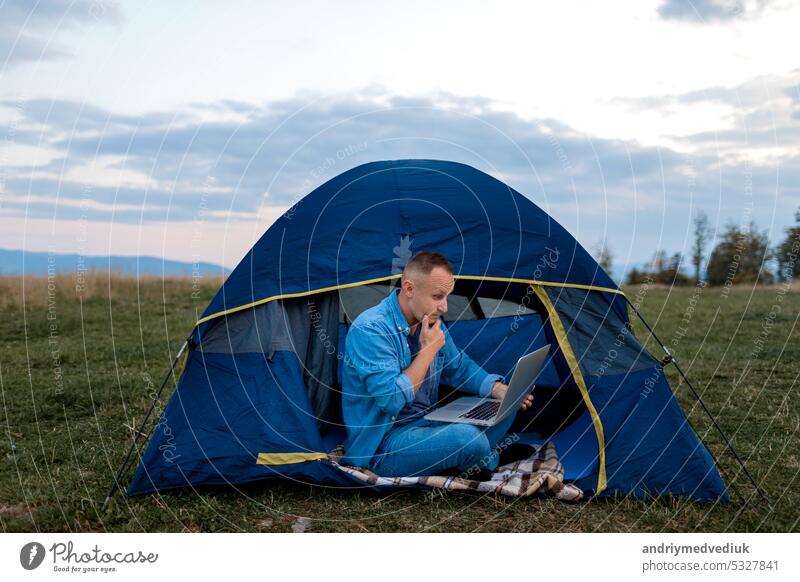 Mann arbeitet am Laptop im Zelt in der Natur. Junger Freiberufler sitzt im Camp. Entspannen in den Bergen. Fernarbeit, Outdoor-Aktivität im Sommer. Glückliche männliche Arbeit im Urlaub.