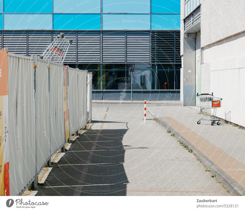 Bauzaun in einem Einkaufszentrum, darauf Einkaufswagen. Straße, dahinter moderne Fassade mit blauen Elementen. shoppen Schatten Sonne Baustelle trostlos Konsum