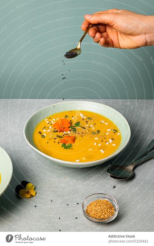 Crop Person, die Sesam in die Kürbiscremesuppe gibt Suppe Sahne Schalen & Schüsseln Samen hinzufügen Gemüse Lebensmittel Tisch Mahlzeit Speise Veganer Möhre