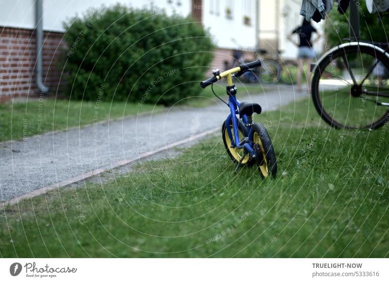 Feierabend! Kinderfahrrad geparkt auf der Wiese vor dem Haus, ein grüner Busch, eine Mama in der Ferne, die ihr spielendes Kind ruft. parken abstellen