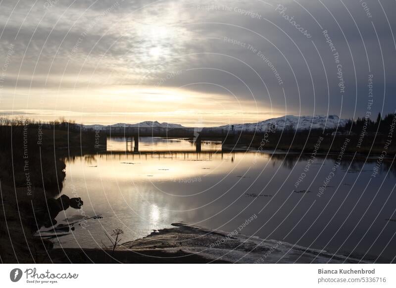 See im Sonnenuntergang im Thingvellir National Park mit der Silhouette einer Brücke und Bergen im Hintergrund. Sonnenuntergangshimmel Himmel gelb