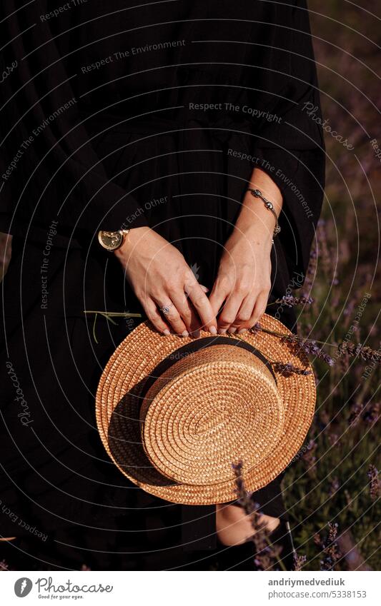 Ausgeschnittenes Foto von Frau in schwarzem Kleid hält Strohhut und Lavendelblume in den Händen in blühenden Lavendelfeld. Mode Schönheit Konzept. Stylish Mädchen mit Schmuck, Uhr auf ihre Hände. Frauentag