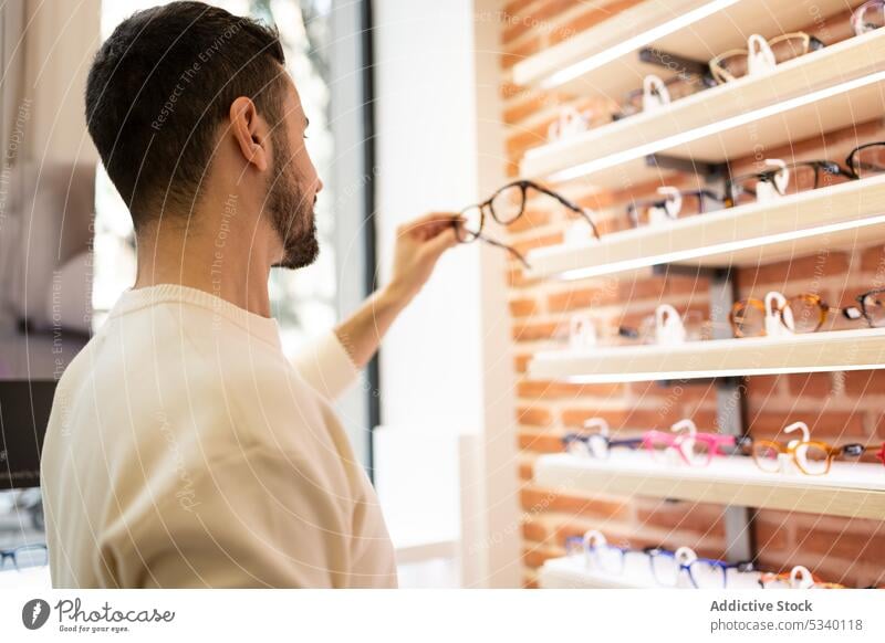 Bärtiger Mann wählt in einem Optikgeschäft eine Brille aus wählen Kunde Werkstatt kaufen Kauf Käufer Laden Regal Klient männlich Wahl Sale modern verbrauchen