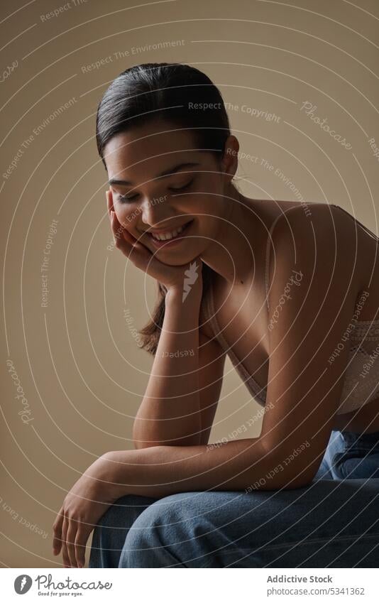 Sinnliche Frau lächelnd mit geschlossenen Augen gegen beige Hintergrund Lächeln Glück Model BH Stil heiter Porträt positiv Augen geschlossen sitzen Jeanshose