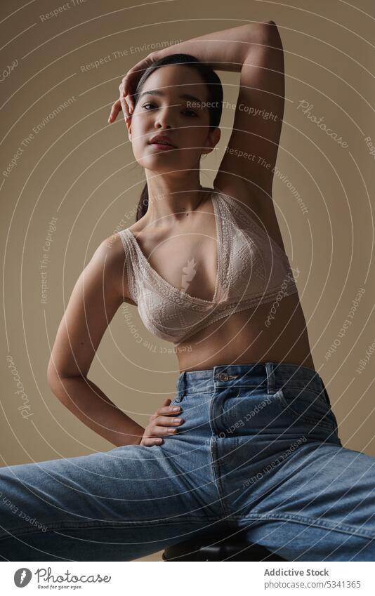 Anmutige Frau sitzt auf einem Stuhl in einem hellen Studio Stil Dessous Porträt Model nachdenklich sinnlich Hocker Jeanshose Unterwäsche Figur Arme hochgezogen