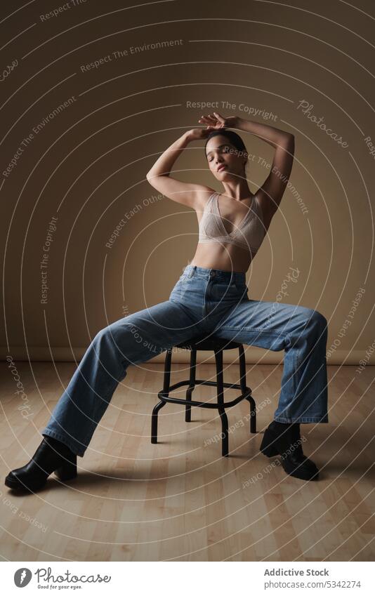 Anmutige Frau sitzt auf einem Stuhl in einem hellen Studio Stil Dessous Porträt Model nachdenklich sinnlich Hocker Jeanshose Augen geschlossen Unterwäsche Figur