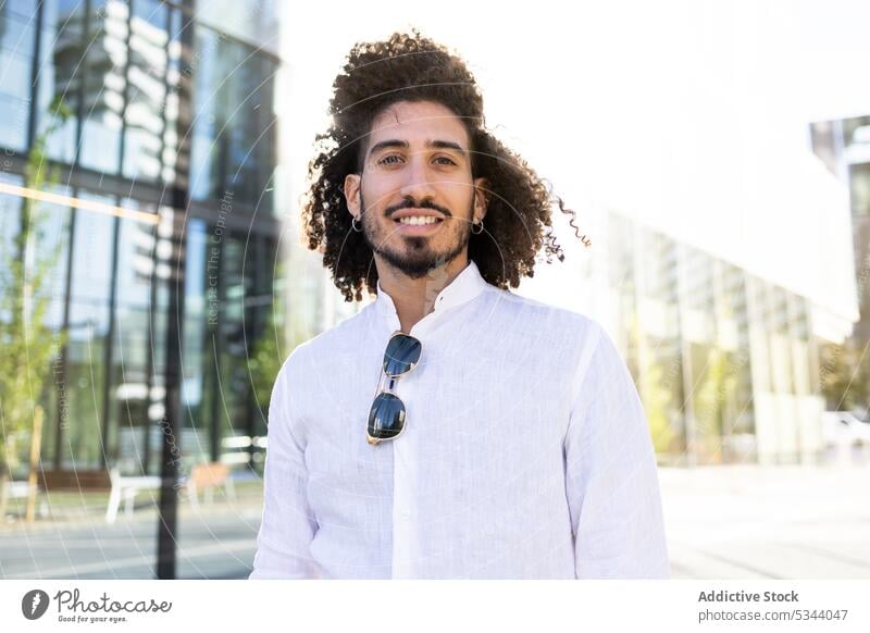 Glücklicher ethnischer Mann steht auf der Straße und schaut in die Kamera positiv Lächeln Stil Großstadt Outfit trendy krause Haare Optimist