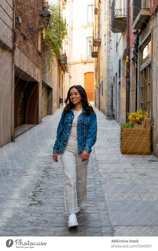 Fröhliche ethnische Frau auf der Straße in der Stadt Gasse Spaziergang schlendern Schatten Gebäude Außenseite eng tagsüber jung Straßenbelag Sommer urban