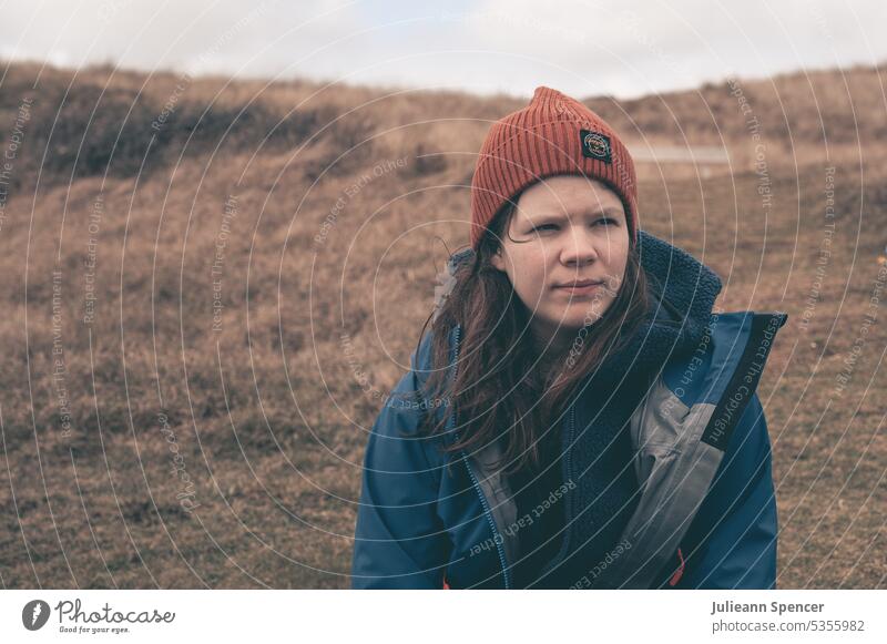 Mädchen mit Mütze und Blick in die Ferne junges Mädchen Beanie Hut wollig warm lange Haare blaue Jacke wasserdichter Mantel kalt Winter totes Gras Sitzen