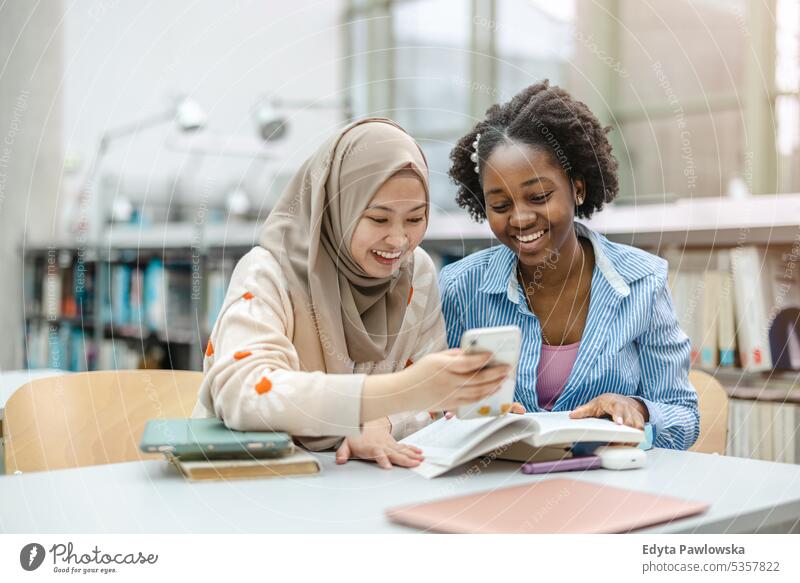 Studentinnen, die in einer Bibliothek sitzen und ihr Smartphone benutzen echte Menschen Teenager Campus positiv Prüfung Wissen selbstbewusst schulisch