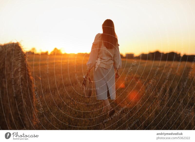 junge frau im schönen licht des sommerlichen sonnenuntergangs auf einem feld geht in der nähe der strohballen. schönes romantisches mädchen mit langen haaren im freien auf dem feld.