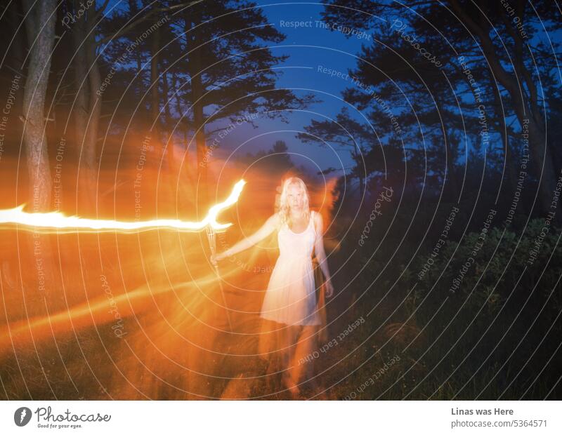 In einer dunklen Sommernacht wandert ein wunderschönes blondes Mädchen in einem weißen Kleid mit einer Fackel herum. Eine Langzeitbelichtung verleiht der Aufnahme eine gewisse Spannung und Unheimlichkeit. Dunkelblauer Himmel, grüne Wälder und eine hübsche Frau in diesem Bild.