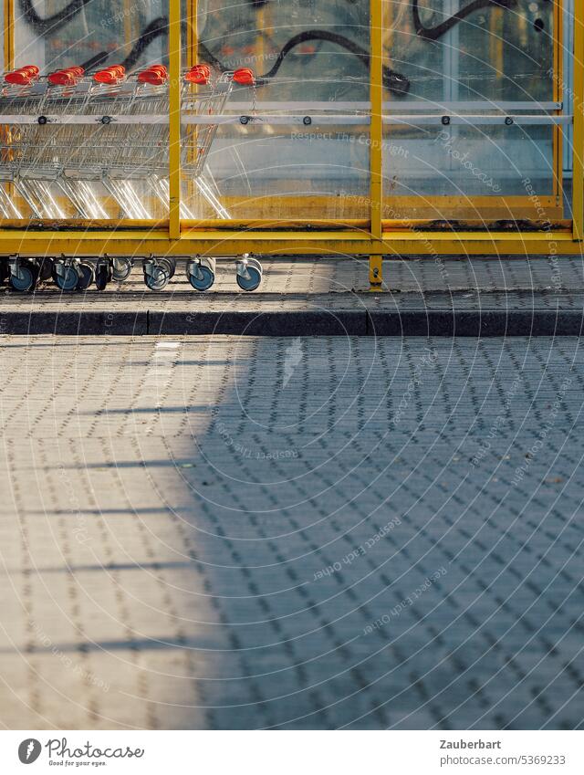 Einkaufswagen in gelber Umhausung, Schatten, gepflasterter Parkplatz Gehäuse Pflaster Pflastersteine Konsum parken kaufen Supermarkt einkaufen Einkaufsmarkt