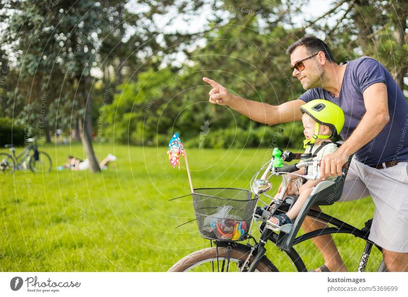 Schauen Sie dort drüben. Aktiver Familientag in der Natur. Vater und Sohn fahren Fahrrad durch den Stadtpark an einem sonnigen Sommertag. Ein niedlicher Junge sitzt im vorderen Fahrradstuhl, während der Vater Fahrrad fährt. Vater-Sohn-Bindung.