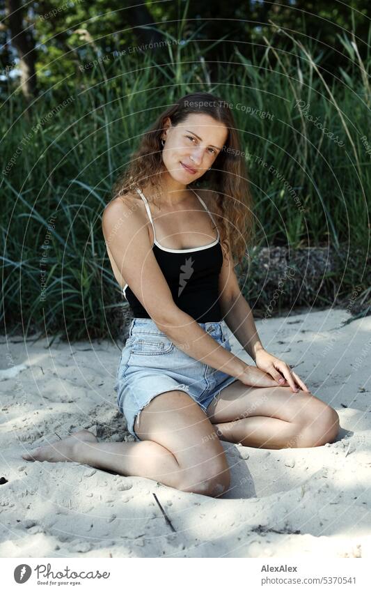 seitliches Portrait einer jungen, schönen, brünetten, lächelnden Frau, die im Sand vor einem Wald kniet warm sommerlich schönes wetter Sommer Pflanzen Nähe Tag