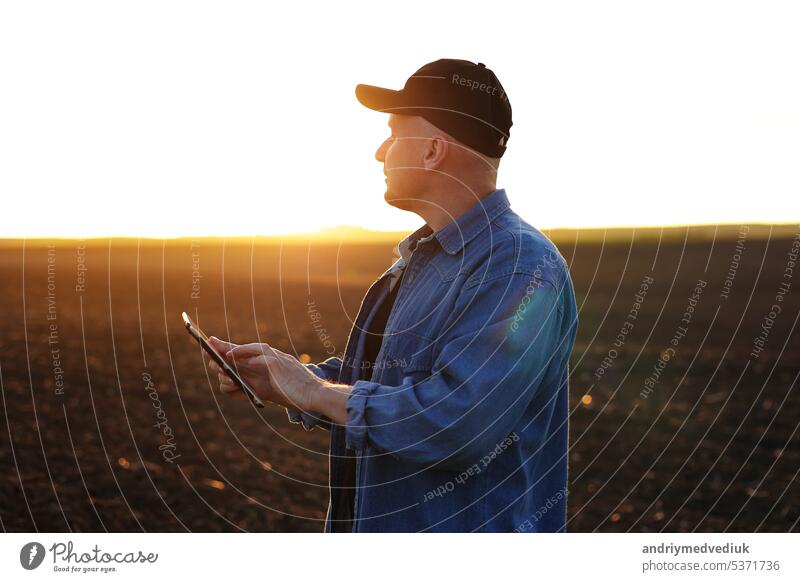 Intelligente Landtechnik und Landwirtschaft. Ein Landwirt benutzt ein digitales Tablet auf einem Feld mit gepflügtem Boden bei Sonnenuntergang. Überprüfung und Kontrolle der Bodenqualität, der Bereitschaft zur Aussaat von Pflanzen und zum Anbau von Gemüse