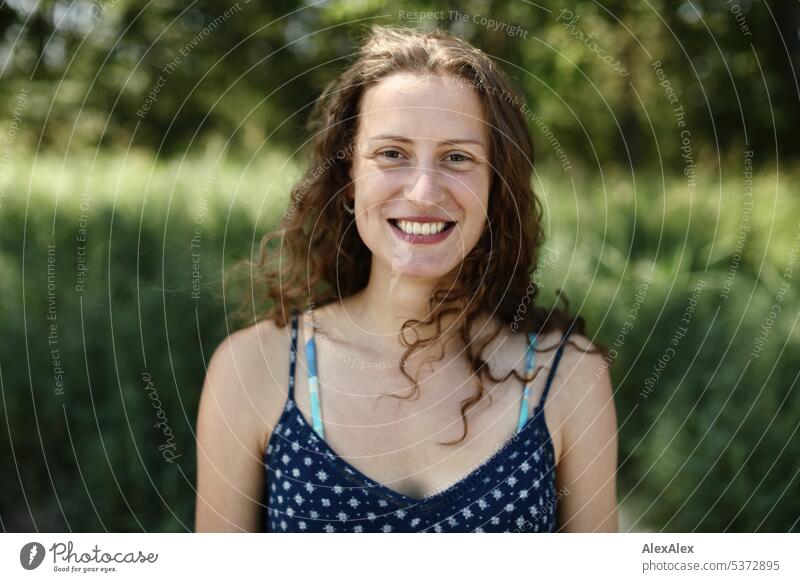 direktes Portrait einer jungen, schönen, brünetten Frau die voller Freude lächelt und vor einem Waldstück steht warm sommerlich schönes wetter Sommer Pflanzen