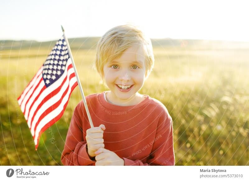 Niedlicher kleiner Junge, der den 4. Juli, den Unabhängigkeitstag der USA, bei sonnigem Sommer-Sonnenuntergang feiert. Kind läuft mit amerikanischer Flagge der Vereinigten Staaten auf Weizenfeld. Stolzer kleiner amerikanischer Junge hält Landesflagge