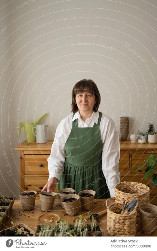 Erwachsene Frau mit Pflanze in Holztopf über Tisch zu Hause Lächeln Gärtner eingetopft Tasse Schürze Gartenbau heimwärts positiv Glück Topf Lebensmitte hölzern