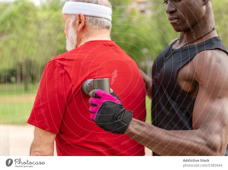 Fit schwarzer Trainer massiert den Rücken eines älteren Kunden mit einem Gerät im Park Männer Massage Trainerin Training Massagegerät Klient Wellness