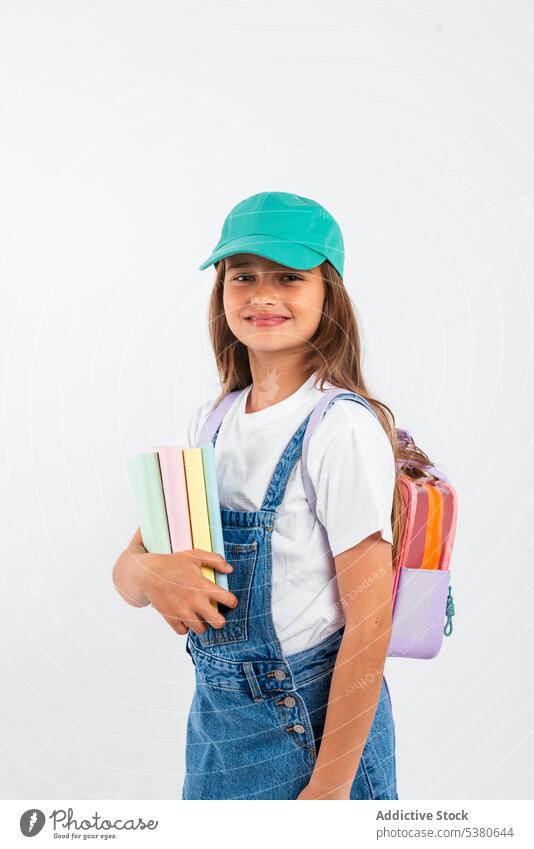 Fröhliches Schulmädchen mit Rucksack Mädchen Lächeln Schule positiv Schüler Kind Bildung Glück Notebook Pupille Jeansstoff Hut führen wenig froh Studioaufnahme