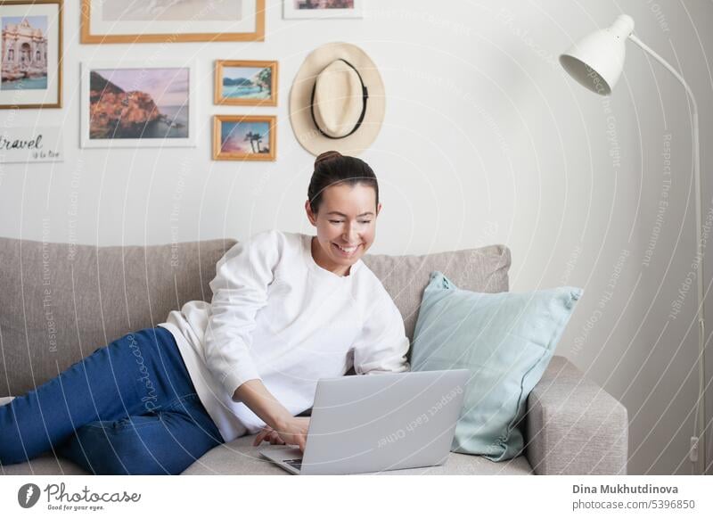 Junge Frau arbeitet am Laptop. Millennial Mädchen arbeiten Remote-Job von zu Hause aus, sitzen auf dem gemütlichen Sofa, lächelnd. Hintergrund schön Business